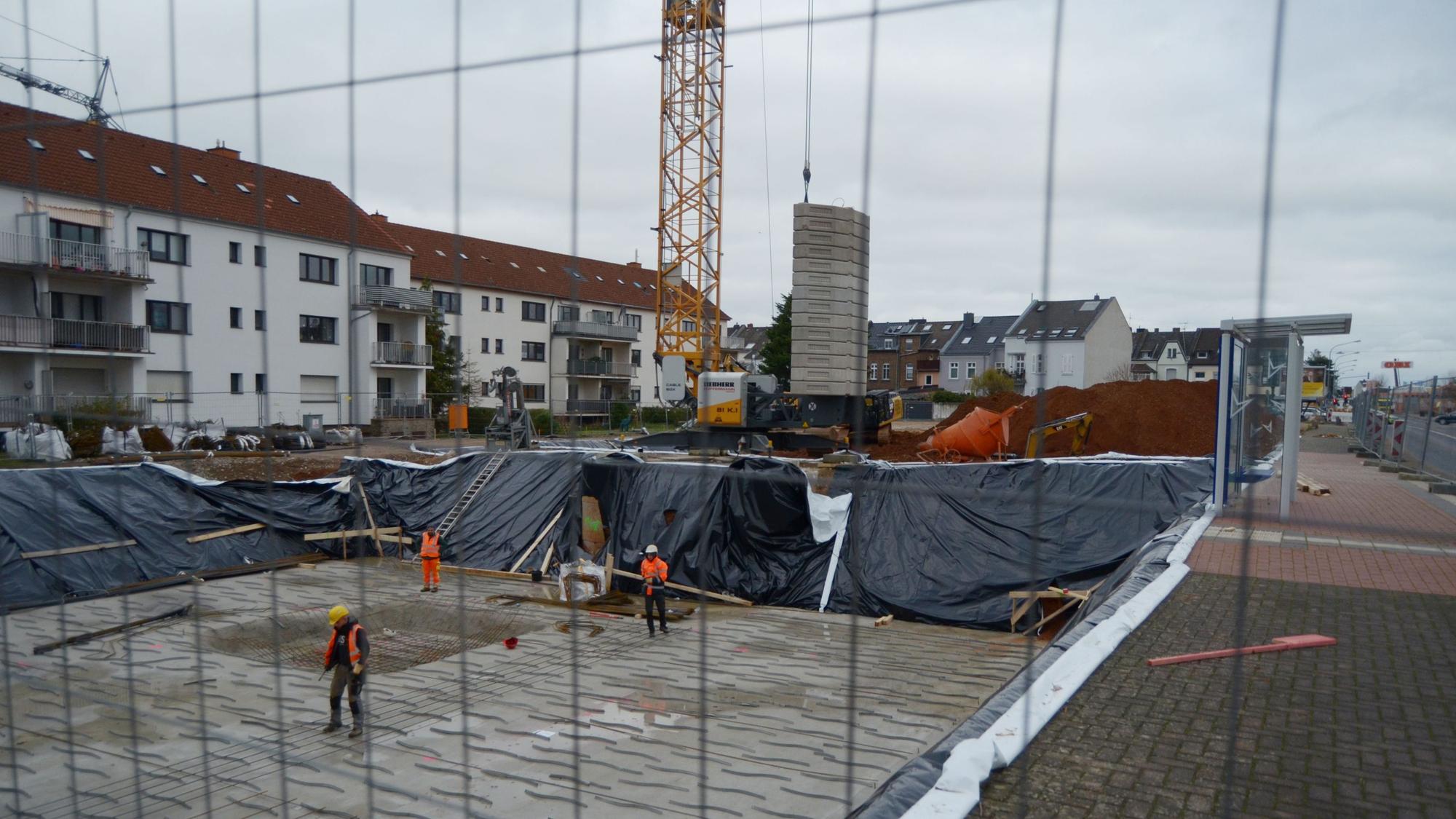 Drei Arbeiter stehen in einer Baugrube, die für die Errichtung eines Mehrfamilienhauses ausgehoben worden ist. Im Hintergrund weitere Wohngebäude und ein Baukran.