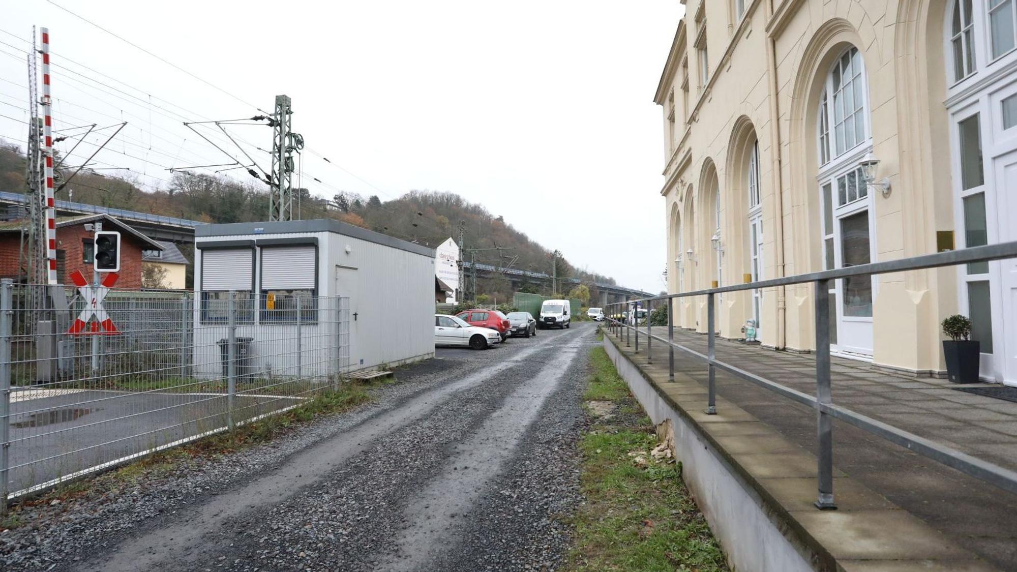 Ein geschotterter Weg führt zwischen dem Bahnübergang Drachenfelsstraße und einem Gebäude entlang.