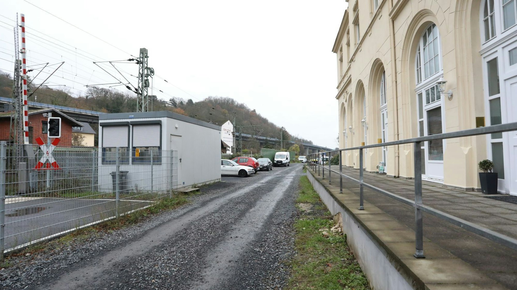Ein geschotterter Weg führt zwischen dem Bahnübergang Drachenfelsstraße und einem Gebäude entlang.