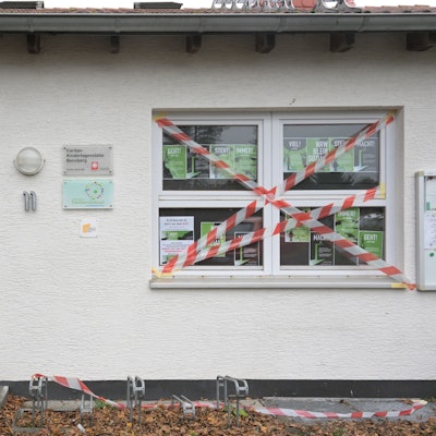 Die Eingangstür und ein Fenster des weißen Kita-Gebäudes sind mit rot-weißem Flatterband beklebt. Im Fenster hängen DinA 4 große Protestplakate.