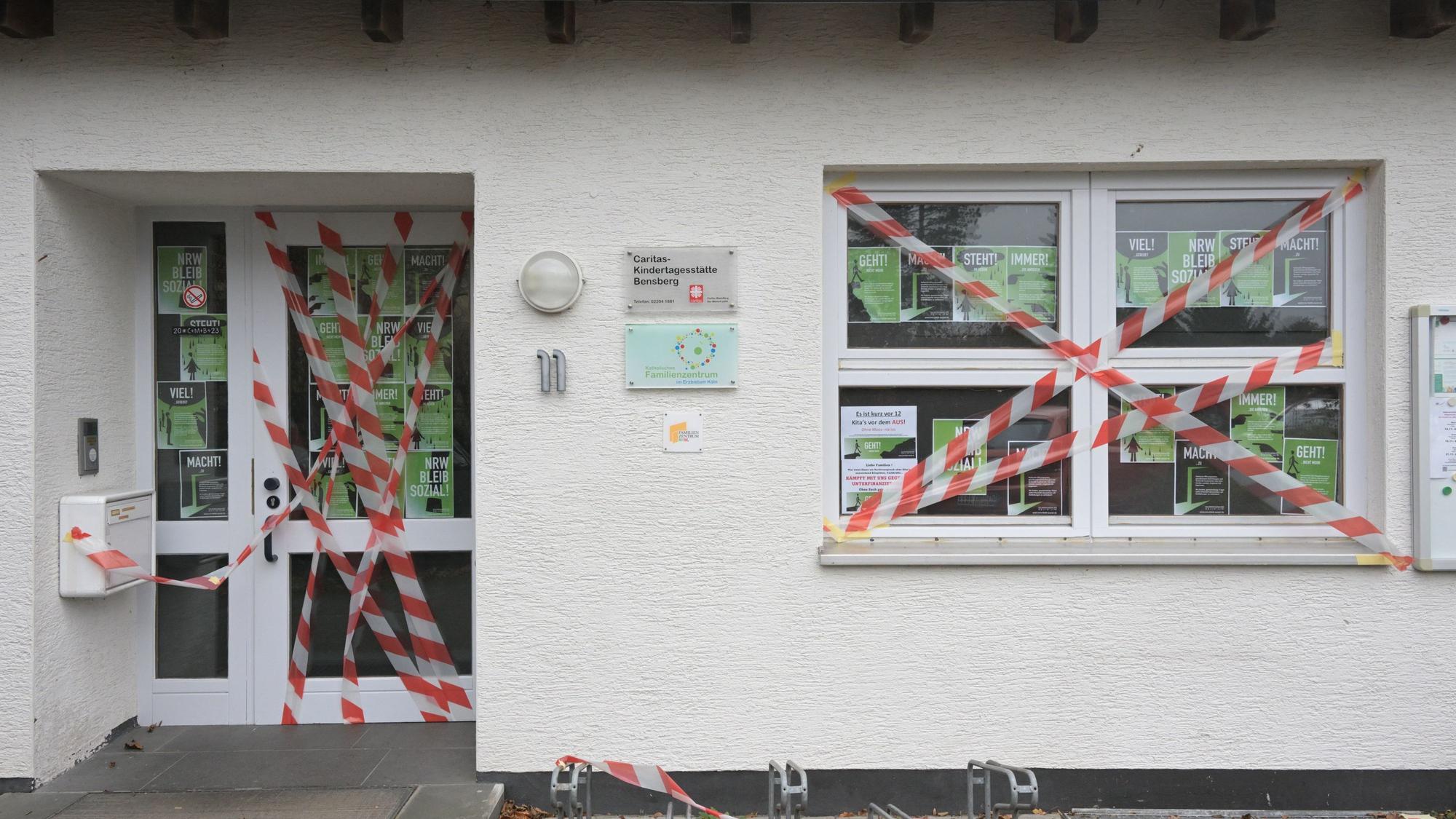 Die Eingangstür und ein Fenster des weißen Kita-Gebäudes sind mit rot-weißem Flatterband beklebt. Im Fenster hängen DinA 4 große Protestplakate.