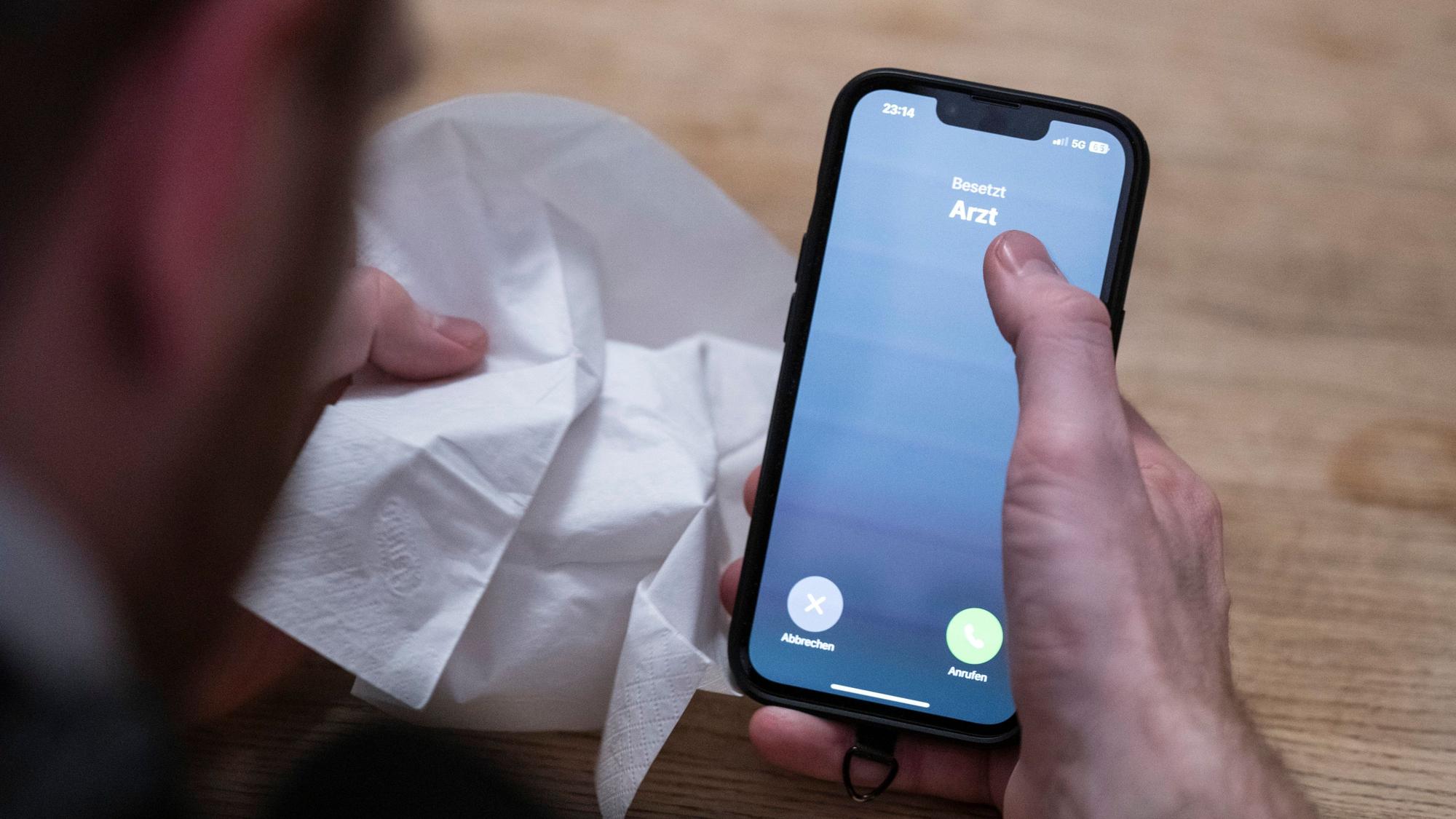 Ein Mann hält ein Taschentuch in einer Hand und ein Telefon mit dem Schriftzug ‚Arzt‘ in der Anderen