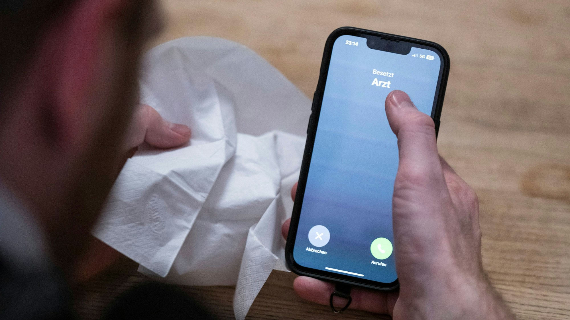 Ein Mann hält ein Taschentuch in einer Hand und ein Telefon mit dem Schriftzug ‚Arzt‘ in der Anderen