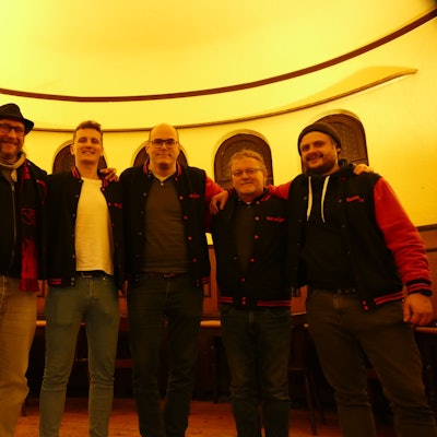 Sechs Männer stehen im Halbkreis in dem Saal einer Kneipe.