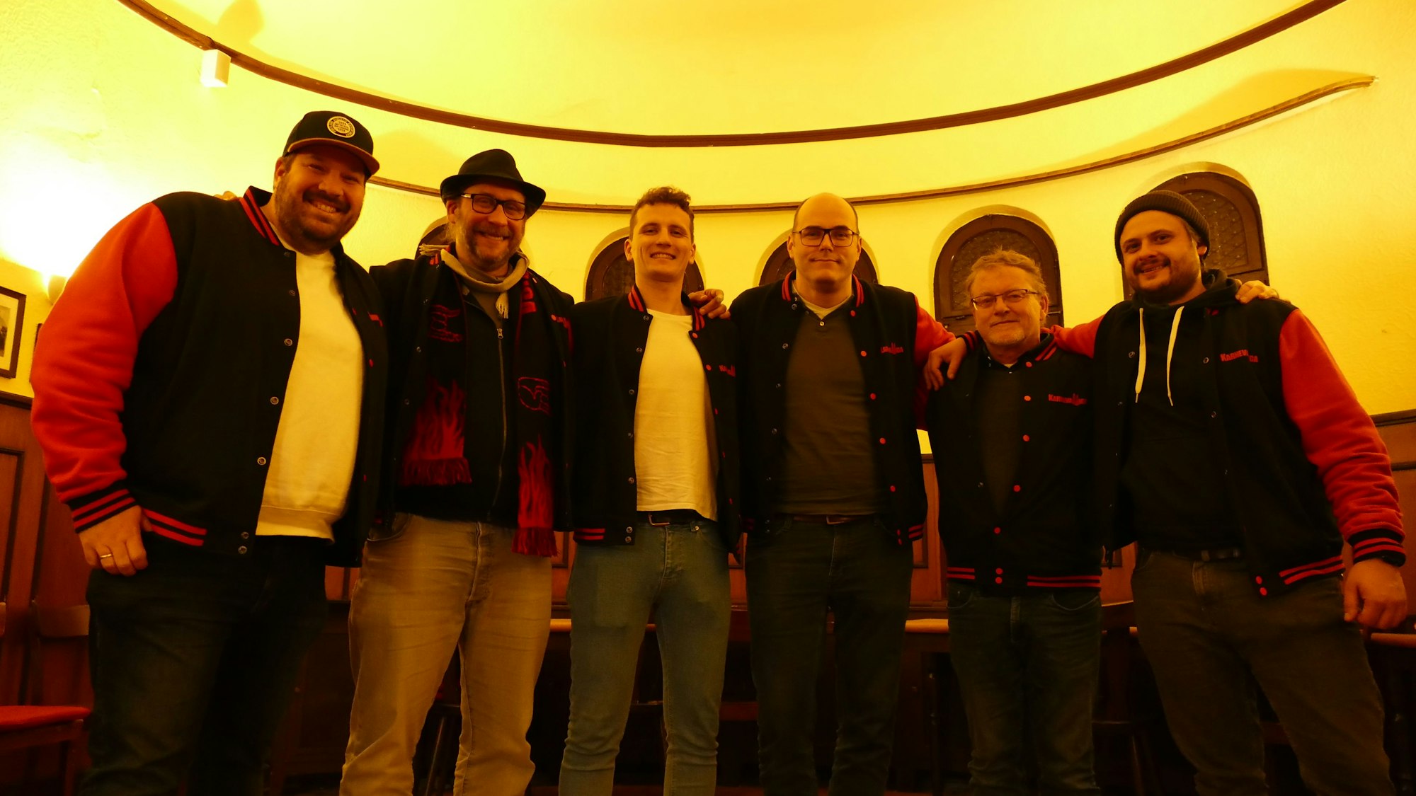 Sechs Männer stehen im Halbkreis in dem Saal einer Kneipe.