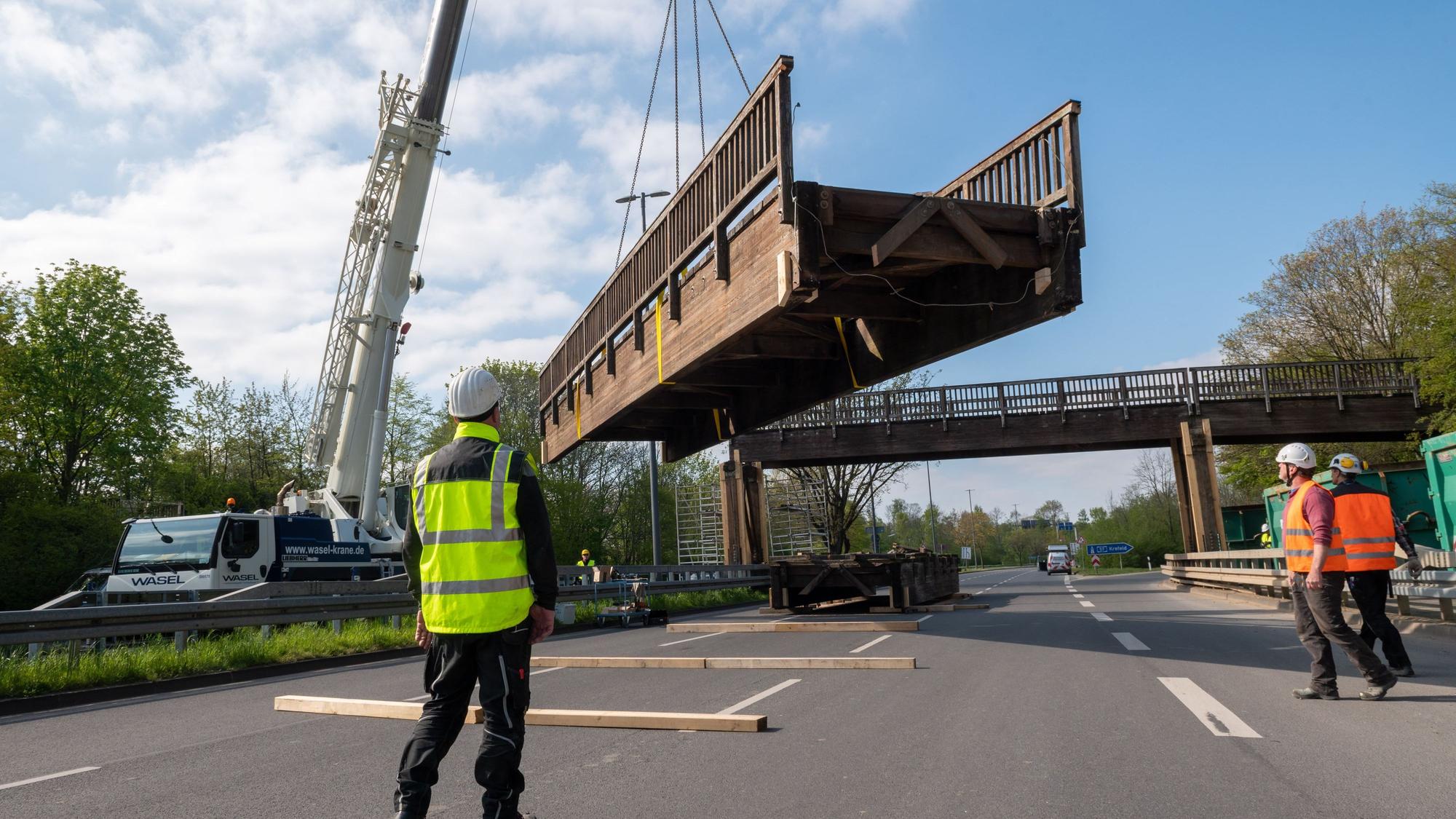 Ein Brückenteil wird von einem Kran angehoben, Arbeiter stehen auf einer mehrspurigen Straße.