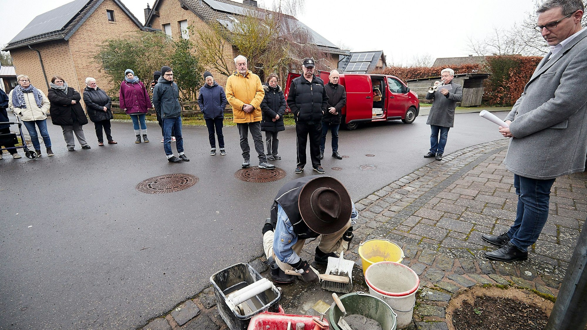 Gunter Demnig kniet auf einem Bürgersteig und verlegt die ersten Stolpersteine in der Gemeinde Nettersheim. Rechts steht Bürgermeister Norbert Crump. Er und einige Anwohner schauen bei der Aktion zu.