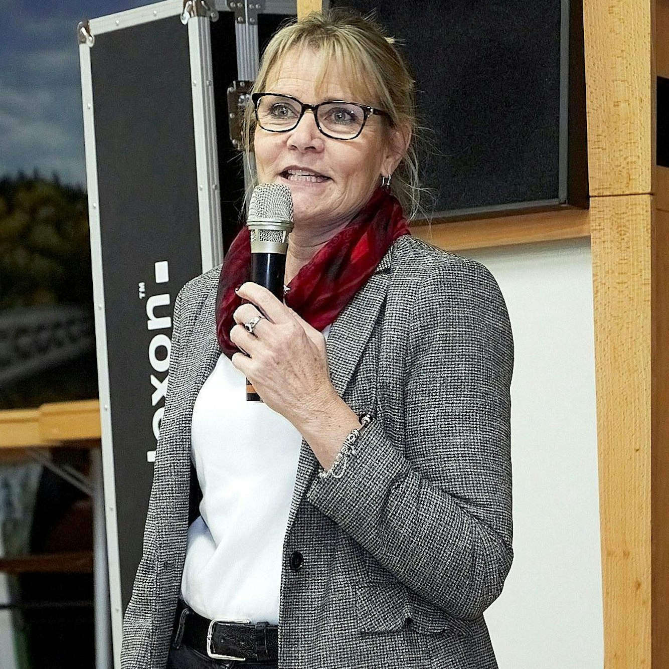 Bärbel König steht vor einer Bühne und spricht in ein Mikrofon. Sie trägt einen grauen Blazer, ein weißes Shirt, schwarze Hose und ein rotes Halstuch. Die blonden Haare hat sie nach hinten gebunden.