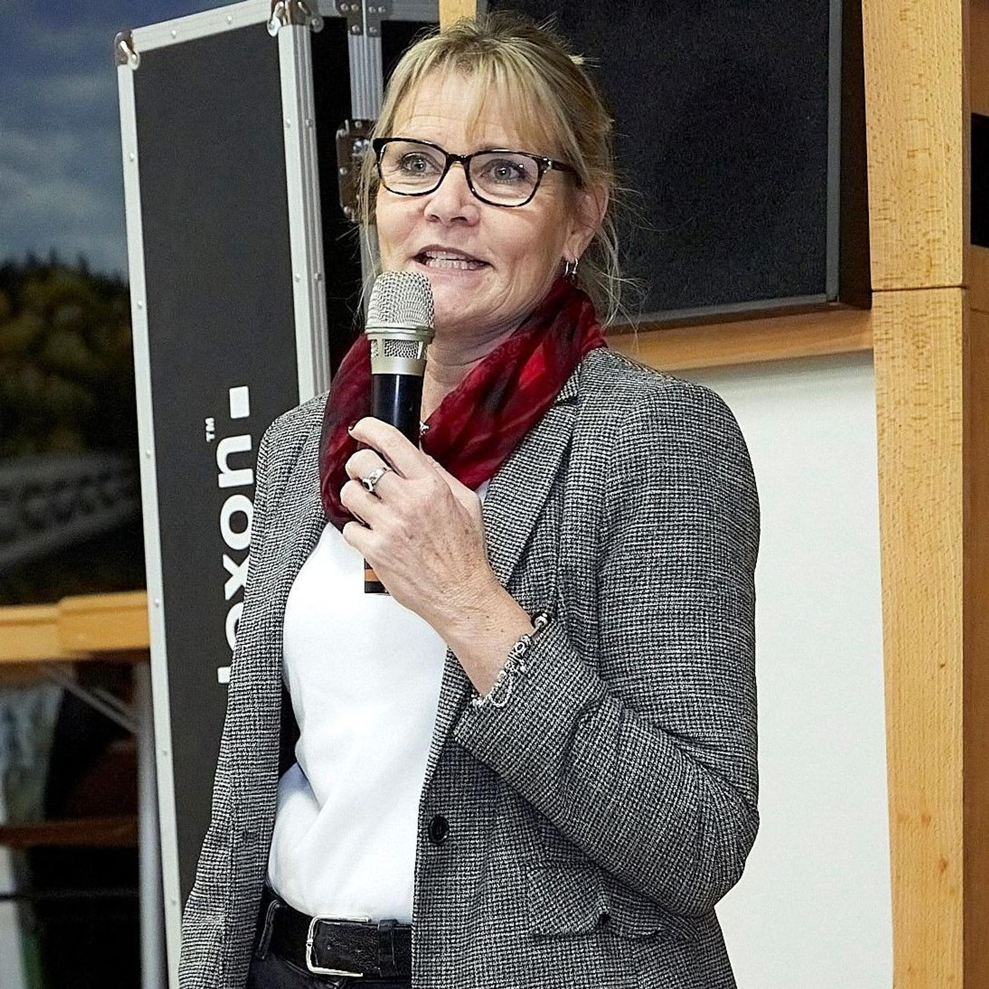 Bärbel König steht vor einer Bühne und spricht in ein Mikrofon. Sie trägt einen grauen Blazer, ein weißes Shirt, schwarze Hose und ein rotes Halstuch. Die blonden Haare hat sie nach hinten gebunden.
