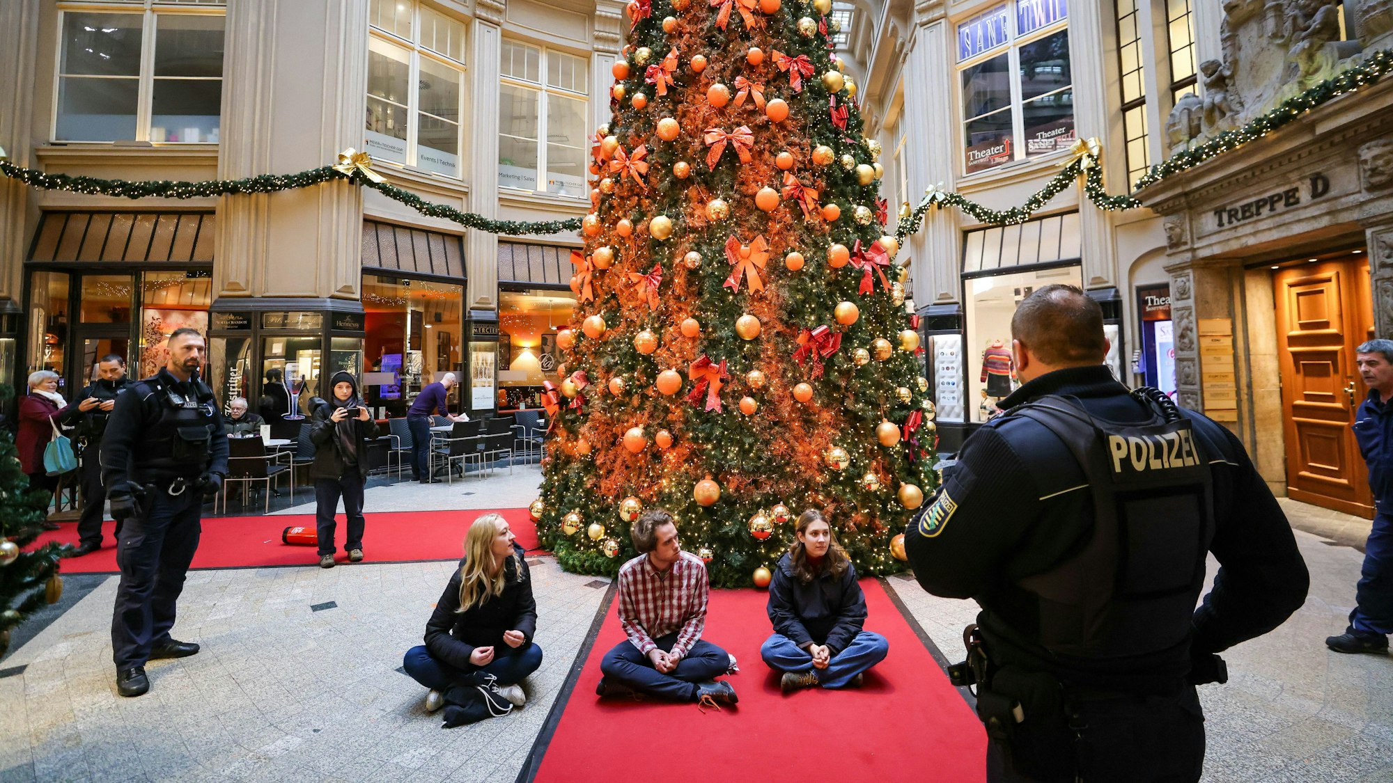 Polizisten stehen um Mitglieder der Protestgruppe Letzte Generation vor einem mit Farbe besprühten Weihnachtsbaum in der Leipziger Mädler-Passage. Die Aktion fand zeitgleich in mehreren Städten statt.