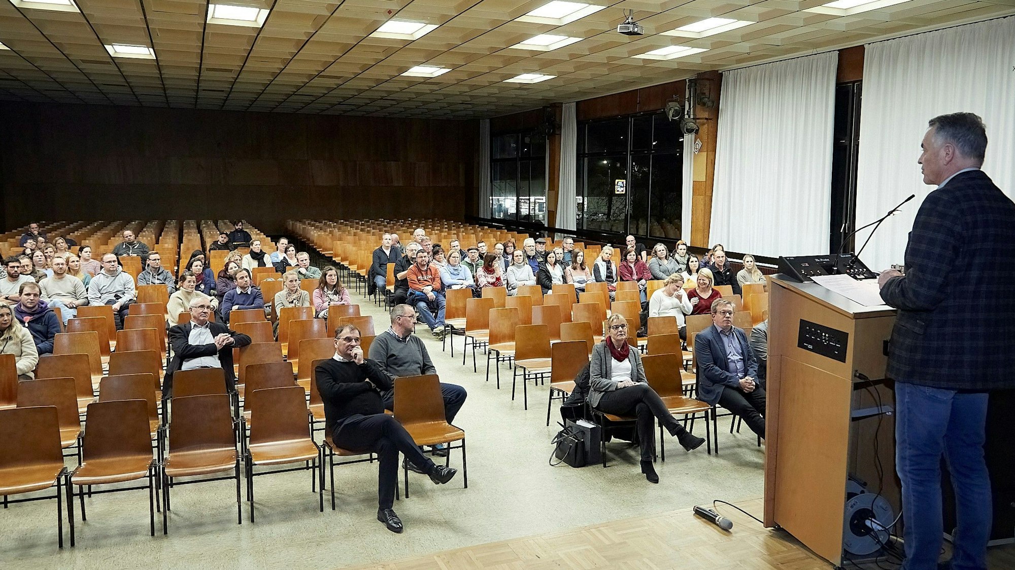 Hellenthals Bürgermeister Rudolf Westerburg steht auf einer Bühne und spricht zu einigen Menschen im Publikum. Viele Stühle sind allerdings leer.