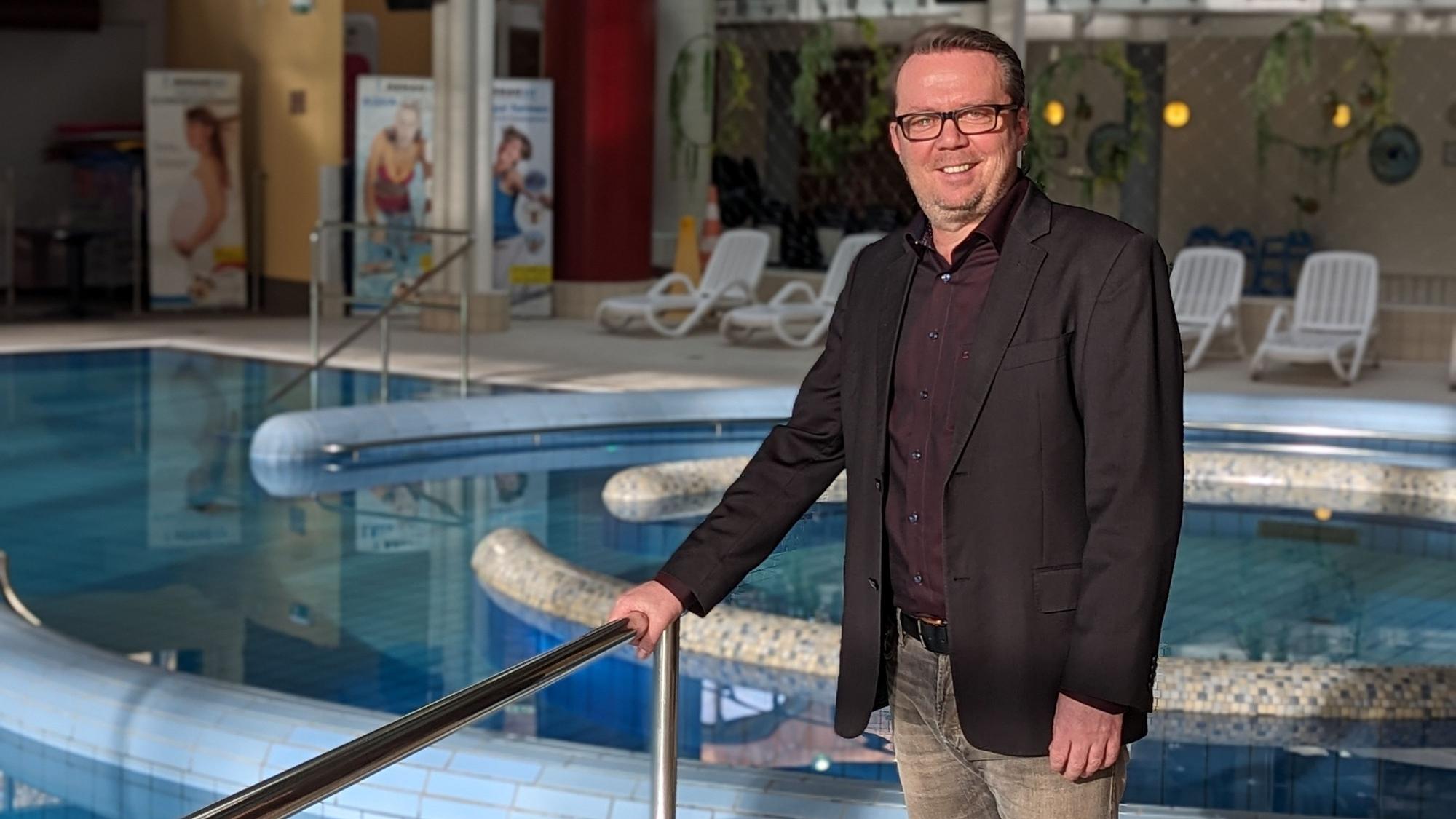 Ein Mann mit dunklem Sakko und Hemd steht vor dem gefüllten Erlebnisbecken im Schwimmbad De Bütt.
