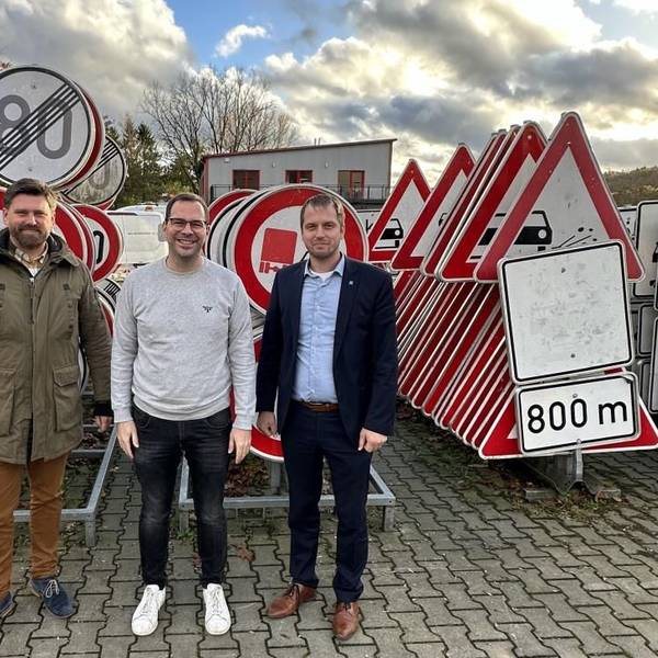 Drei Männer stehen vor einer großen Zahl aufgereihter Verkehrsschilder.