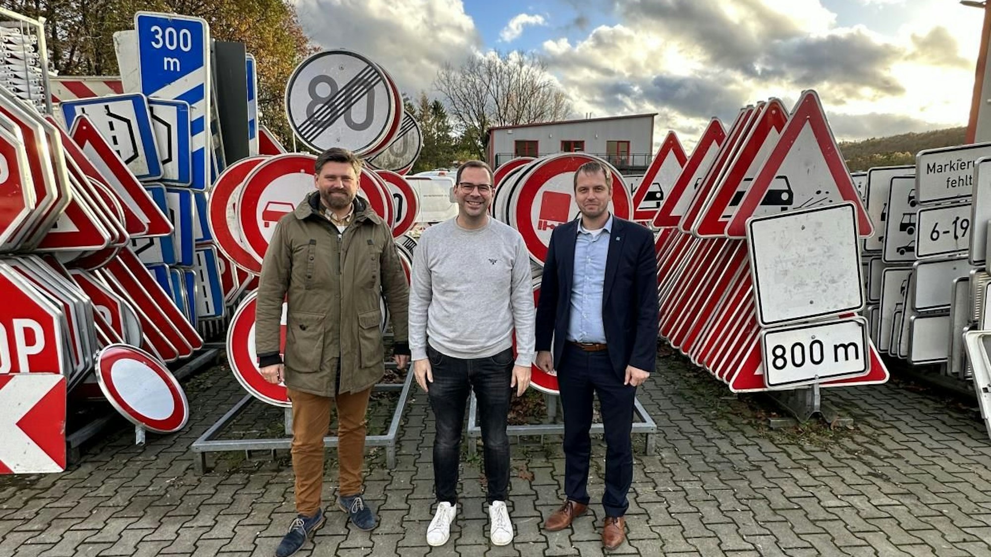 Drei Männer stehen vor einer großen Zahl aufgereihter Verkehrsschilder.