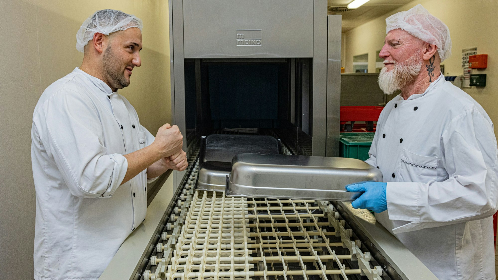 Die beiden stehen sich in weißer Arbeitskleidung gegenüber am Spülmaschinenband.