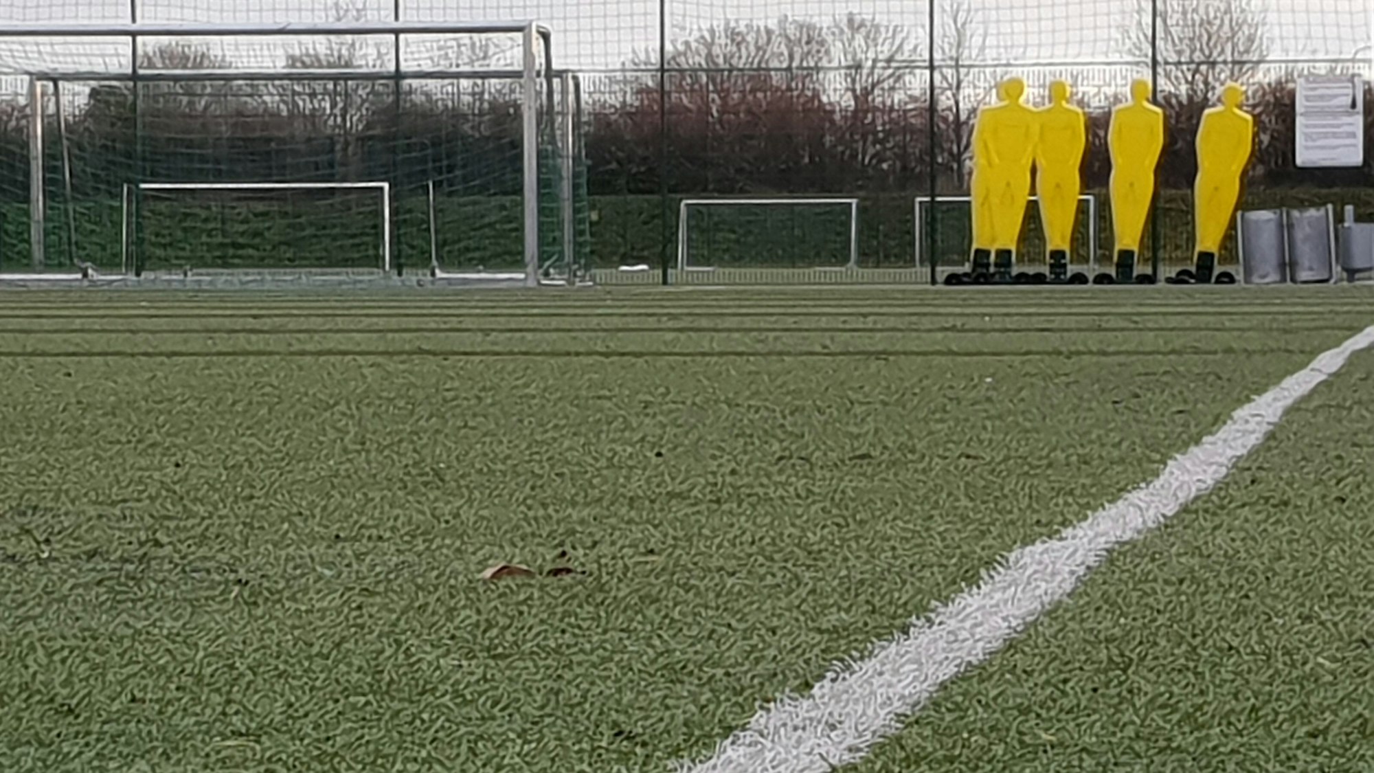 Ein Kunstrasenplatz mit weißer Seitenlinie, Figuren und Toren im Hintergrund.