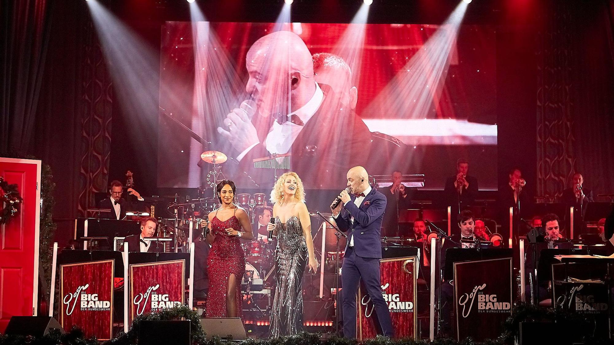 Im Vordergrund der Bühne sind Sänger Marco Matias und die Sängerinnen Sofia Andersson und Tertia Botha zu sehen, dahinter die Musiker in Uniform. Eine Großleinwand dahinter zeigt Sänger Marco Matias.