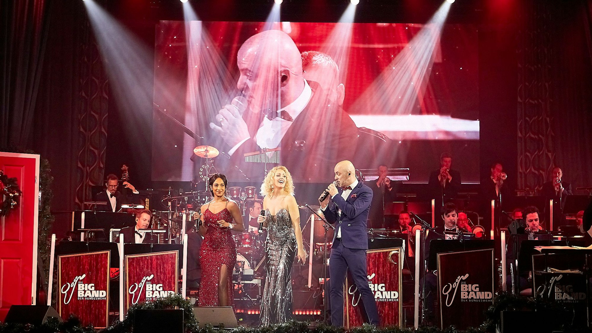 Im Vordergrund der Bühne sind Sänger Marco Matias und die Sängerinnen Sofia Andersson und Tertia Botha zu sehen, dahinter die Musiker in Uniform. Eine Großleinwand dahinter zeigt Sänger Marco Matias.