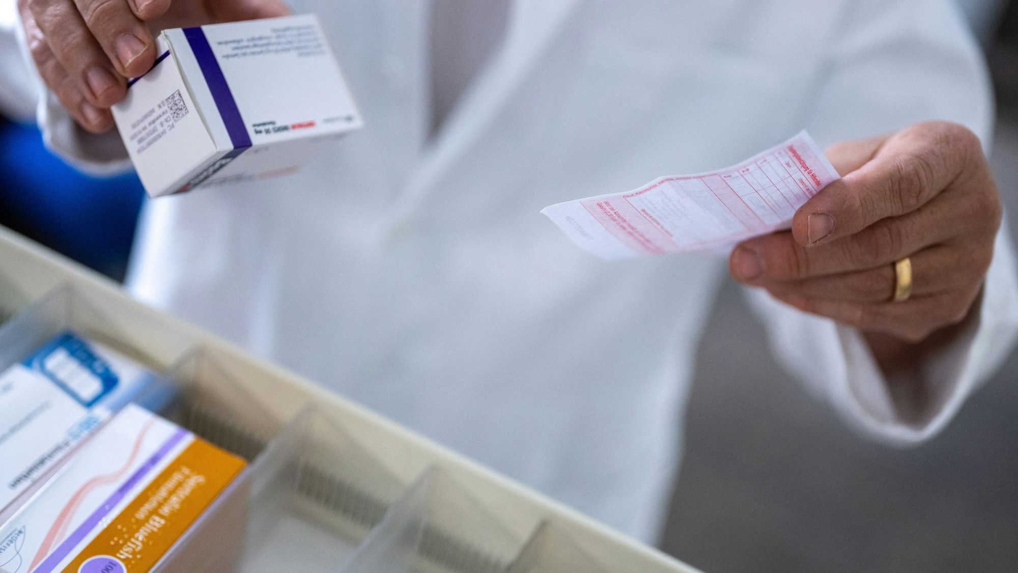 Ein Apotheker hält ein Rezept in der Hand, während er eine Medikamentenverpackung aus einer Schublade in einer Apotheke holt.