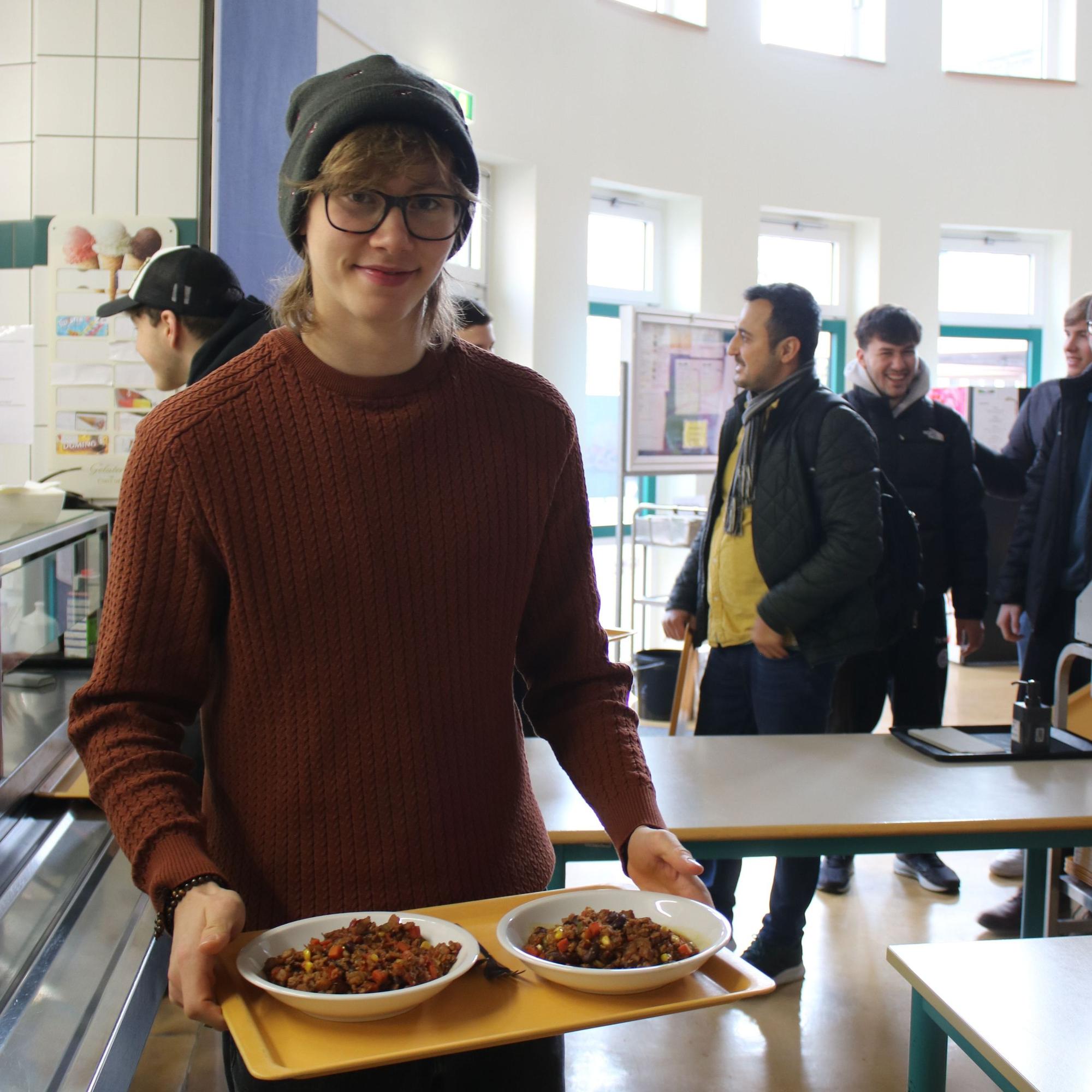 Ein junger Mann steht mit einem Tablett und Essen in einer Schulmensa.