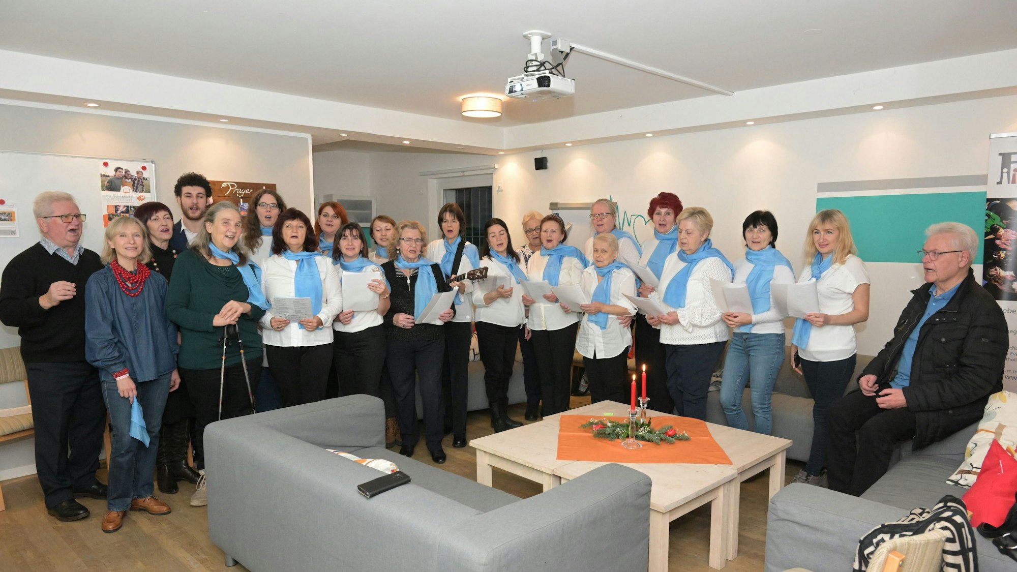 Frauen stehen in weißen T-Shirts und mit blauen Schals in einem Raum und halten Notenblätter.