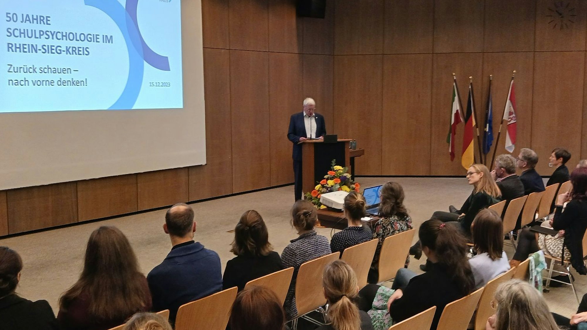 Feiergäste sitzen auf Stühlen in einem Sitzungsaal, ein Mann steht am Rednerpult.