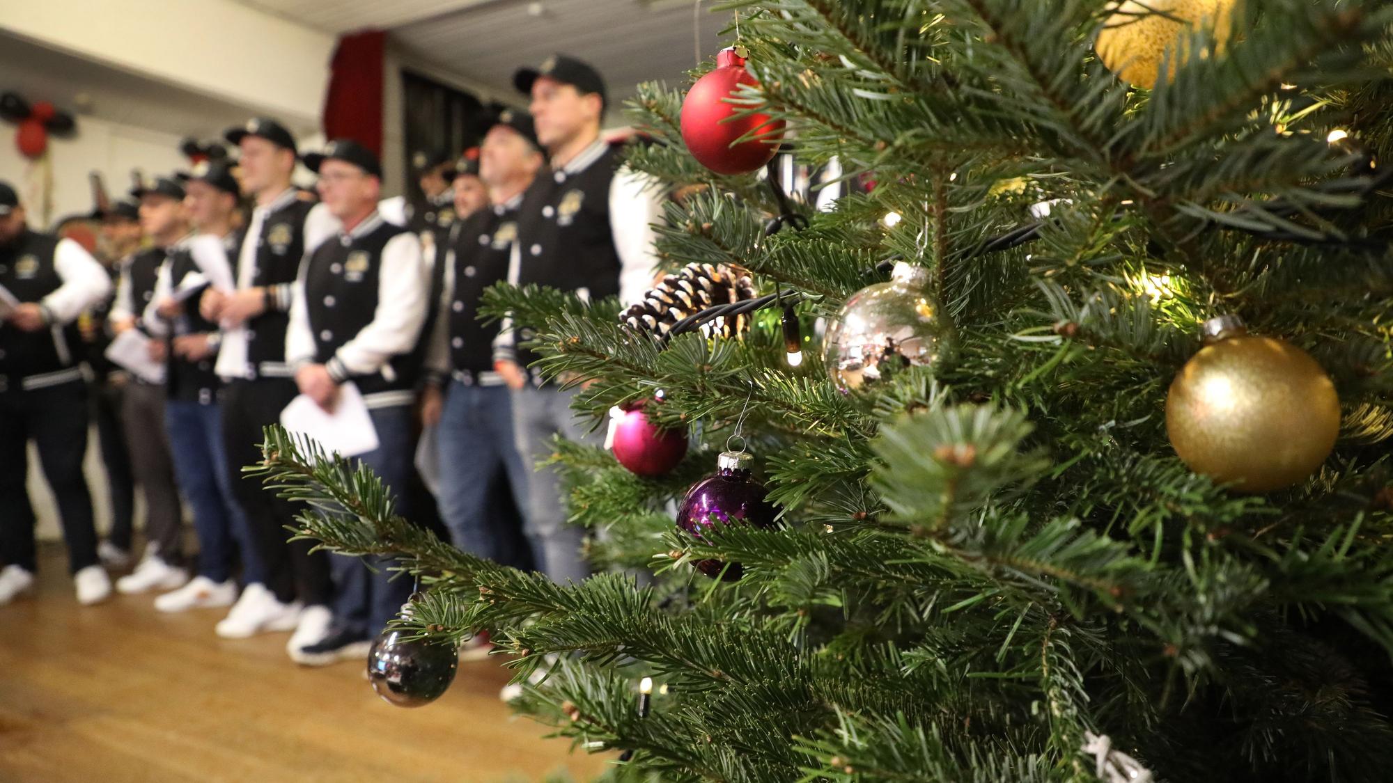 Ein Weihnachtsbaum steht auf einer Bühne, daneben steht ein Männerchor uns singt.