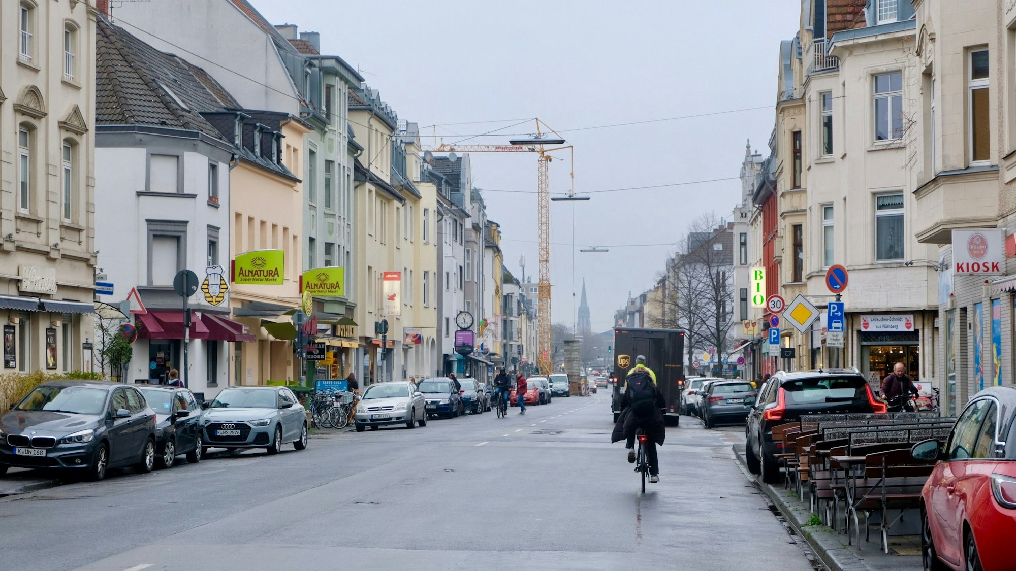 Eine Straße mit parkenden Autos am Rand und Radfahrern ist zu sehen.