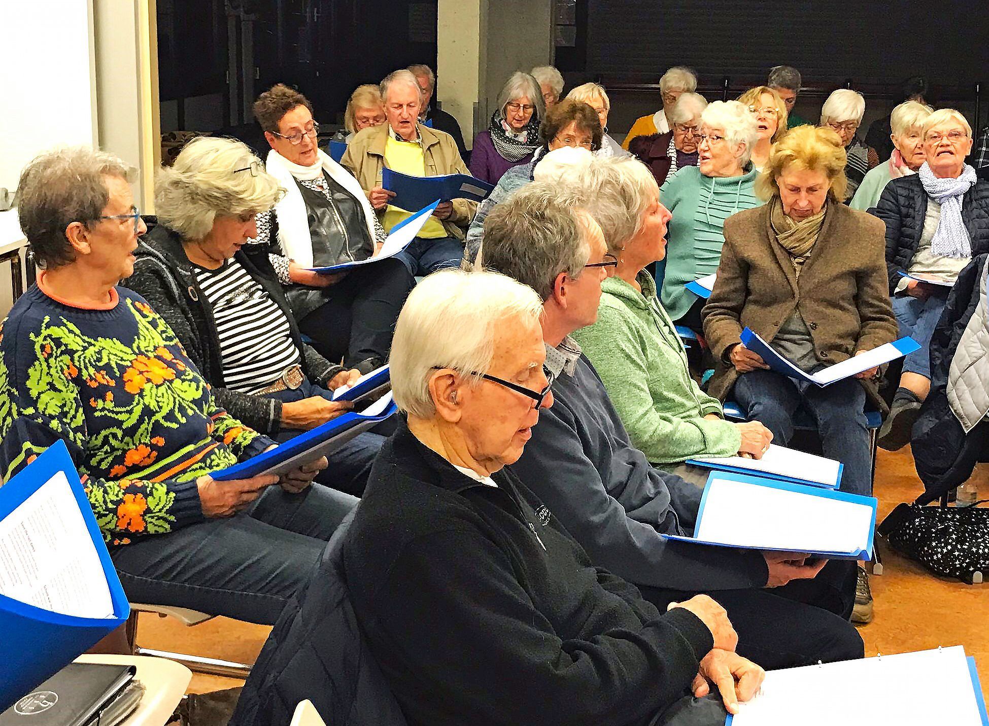 Menschen sitzen nebeneinander und singen, mit Liedmappen auf den Knien.