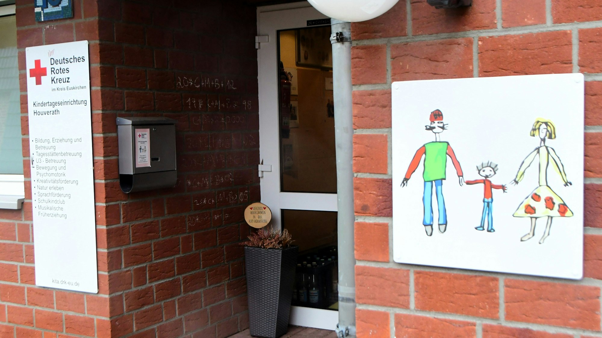 Außenansicht des Eingangsbereichs der Kita Houverath. Rechts ein gezeichnetes Schild mit Mutter, Vater und Kind, links das des Trägers Deutsches Rotes Kreuz.