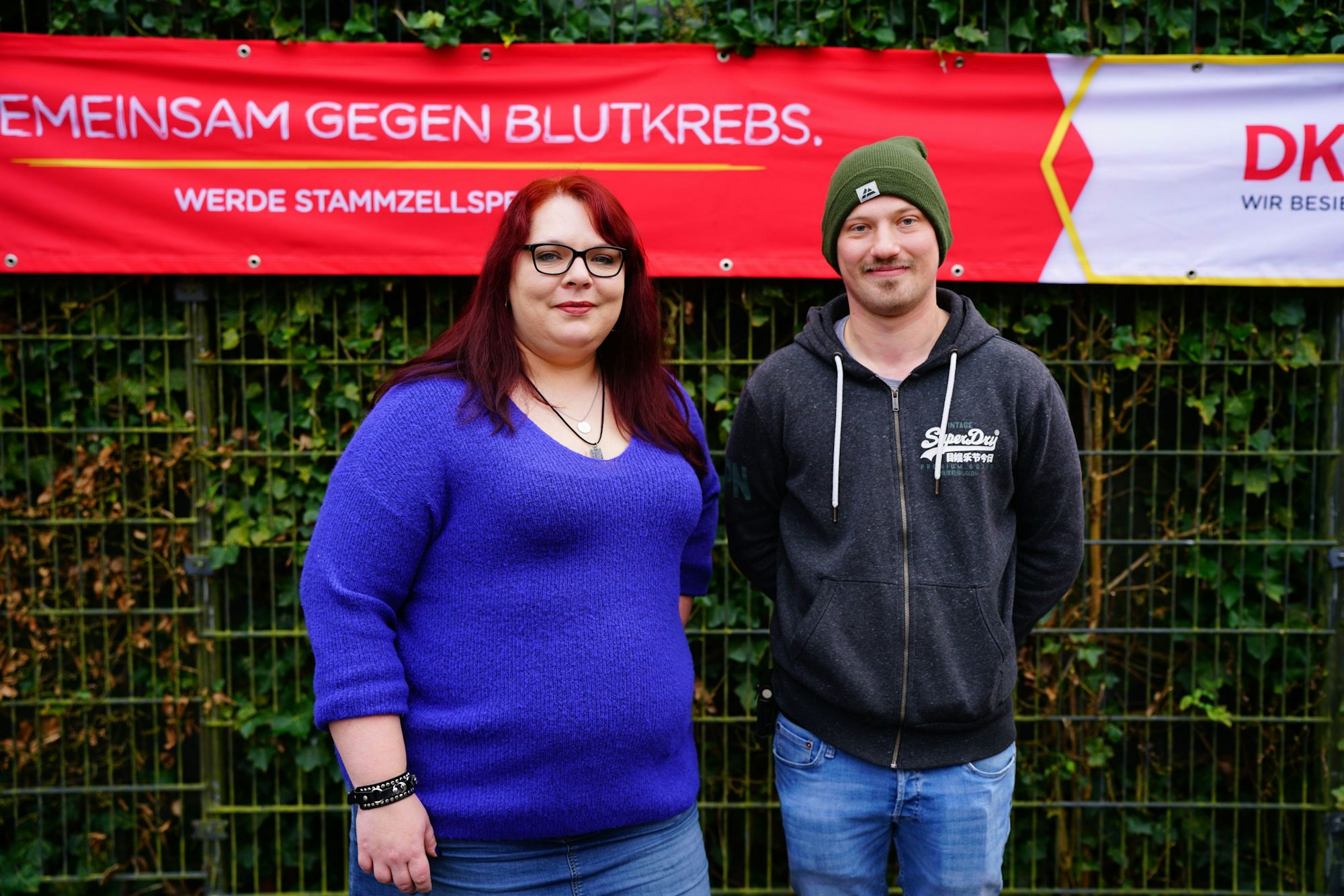 Eine Frau und ein Mann vor einem Zaun mit einem rot-weißen Werbebanner „gemeinsam gegen Blutkrebs“ - werde Stammzellenspender.