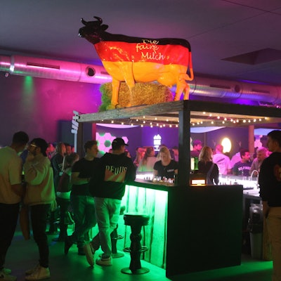 Menschen stehen rund um eine Theke, auf der eine beleuchtete Kuh-Skulptur steht.