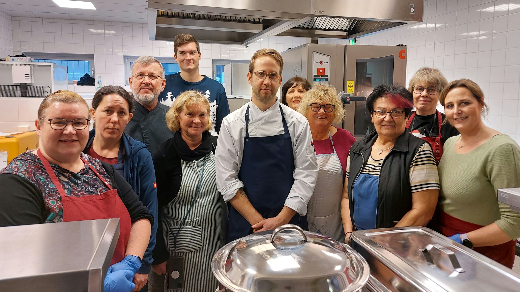 Männer und Frauen stehen in einer Großküche; in der Mitte ein Mann mit Schürze und Kochjacke. Im Vordergrund stehen große Töpfe aus Edelstahl.