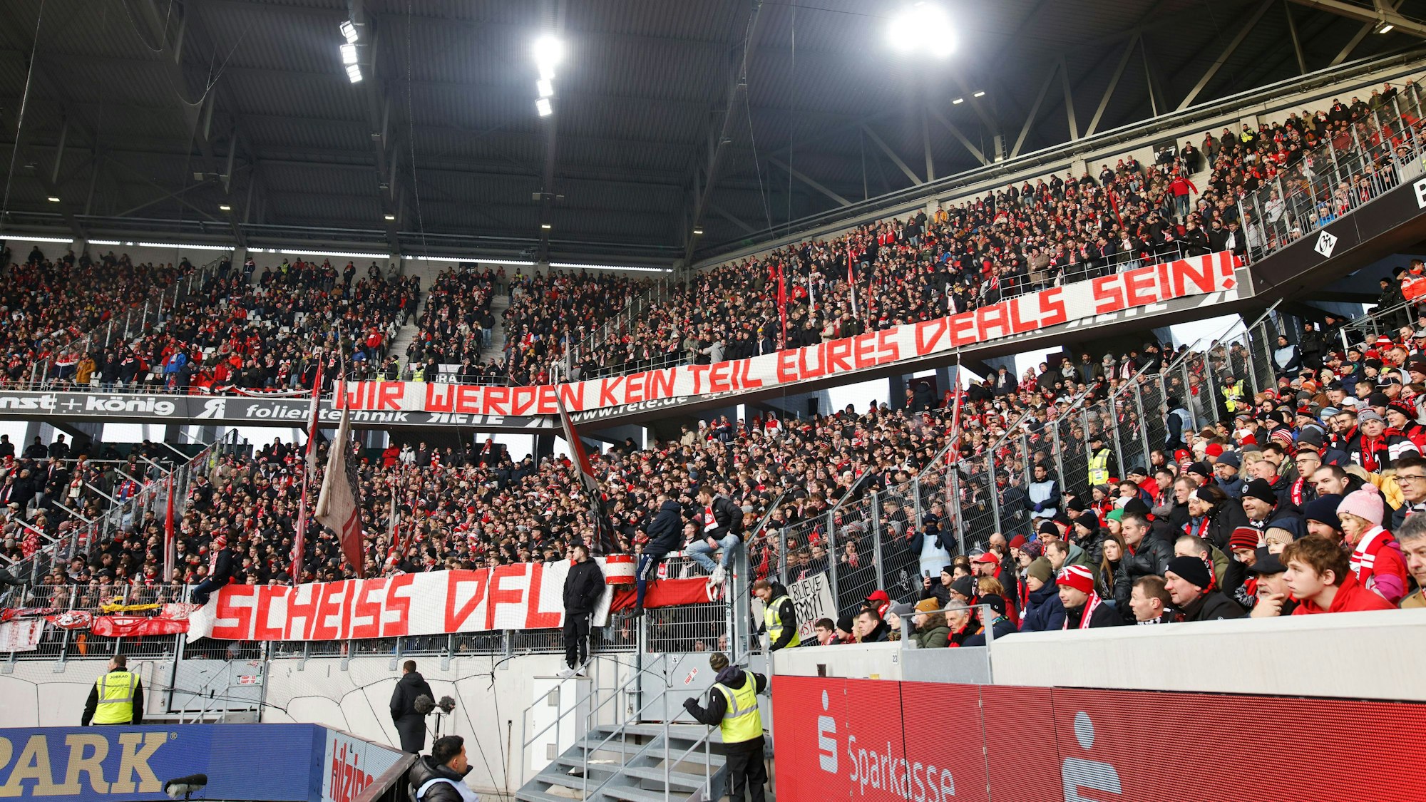 Kölner Fans zeigen Banner mit den Aufschriften „Wir werden kein Teil eures Deals sein!“ und „Scheiss DFL!“ im Gästeblock.