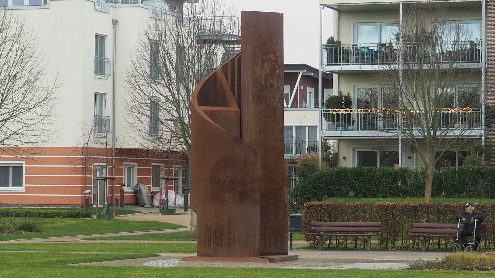 Eine rostige Stahlskulptur in einem Park mit Bänken vor Häusern.