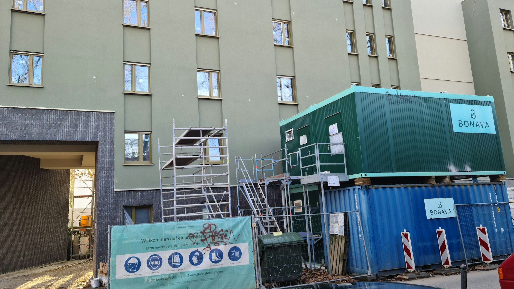 Vor einem Wohngebäude stehen ein Gerüst und Baucontainer.