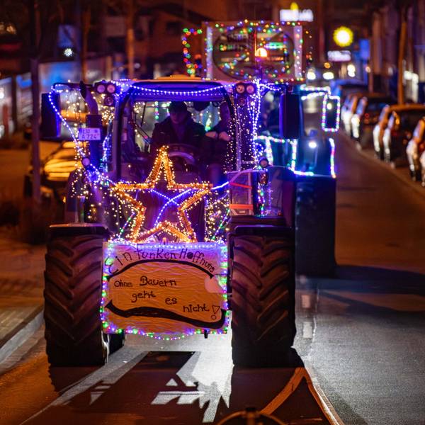 Mehre Traktoren fahren beleuchtet und mit Protestschildern versehen durch die Straßen. Auf einem steht: „Ohne Bauern geht es nicht“
