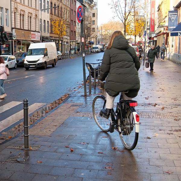 Eine Frau und eine Radfahrerin queren die Bonner Straße auf einem Zebrastreifen, vor dem ein Bus wartet.
