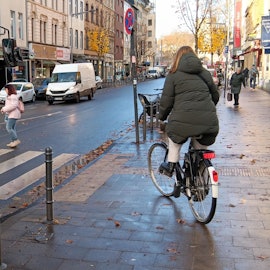 Eine Frau und eine Radfahrerin queren die Bonner Straße auf einem Zebrastreifen, vor dem ein Bus wartet.