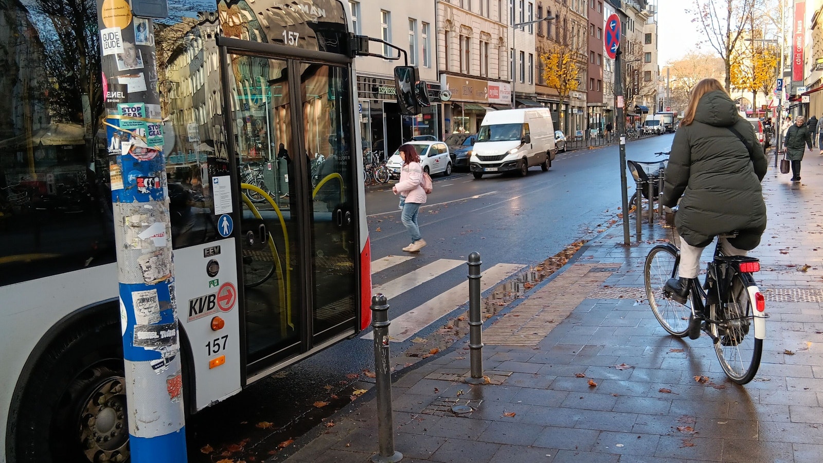Eine Frau und eine Radfahrerin queren die Bonner Straße auf einem Zebrastreifen, vor dem ein Bus wartet.