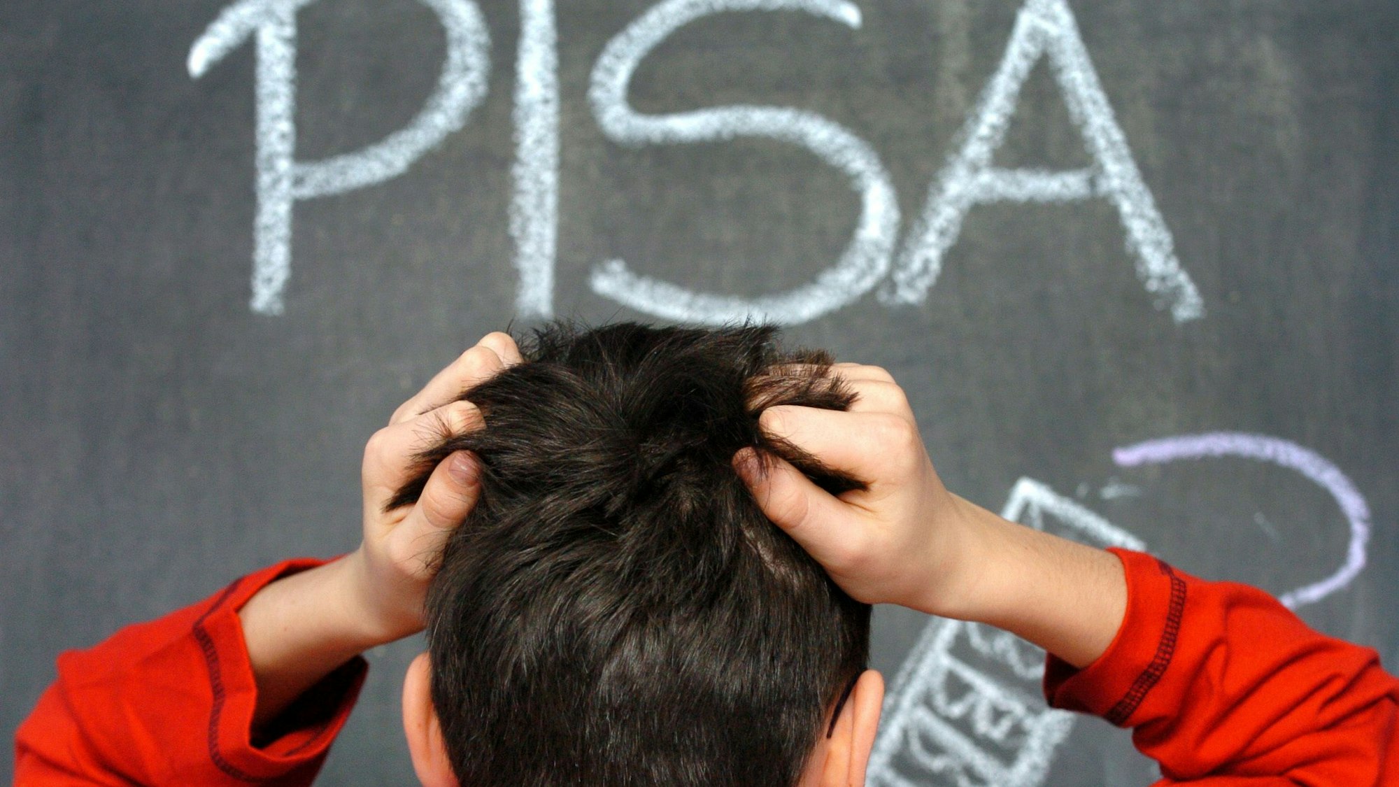 Ein Junge rauft sich die Haare. Im Hintergrund ist eine grüne Tafel in einem Klassenzimmer zu sehen. Auf dieser steht mit Kreide geschrieben: Pisa.