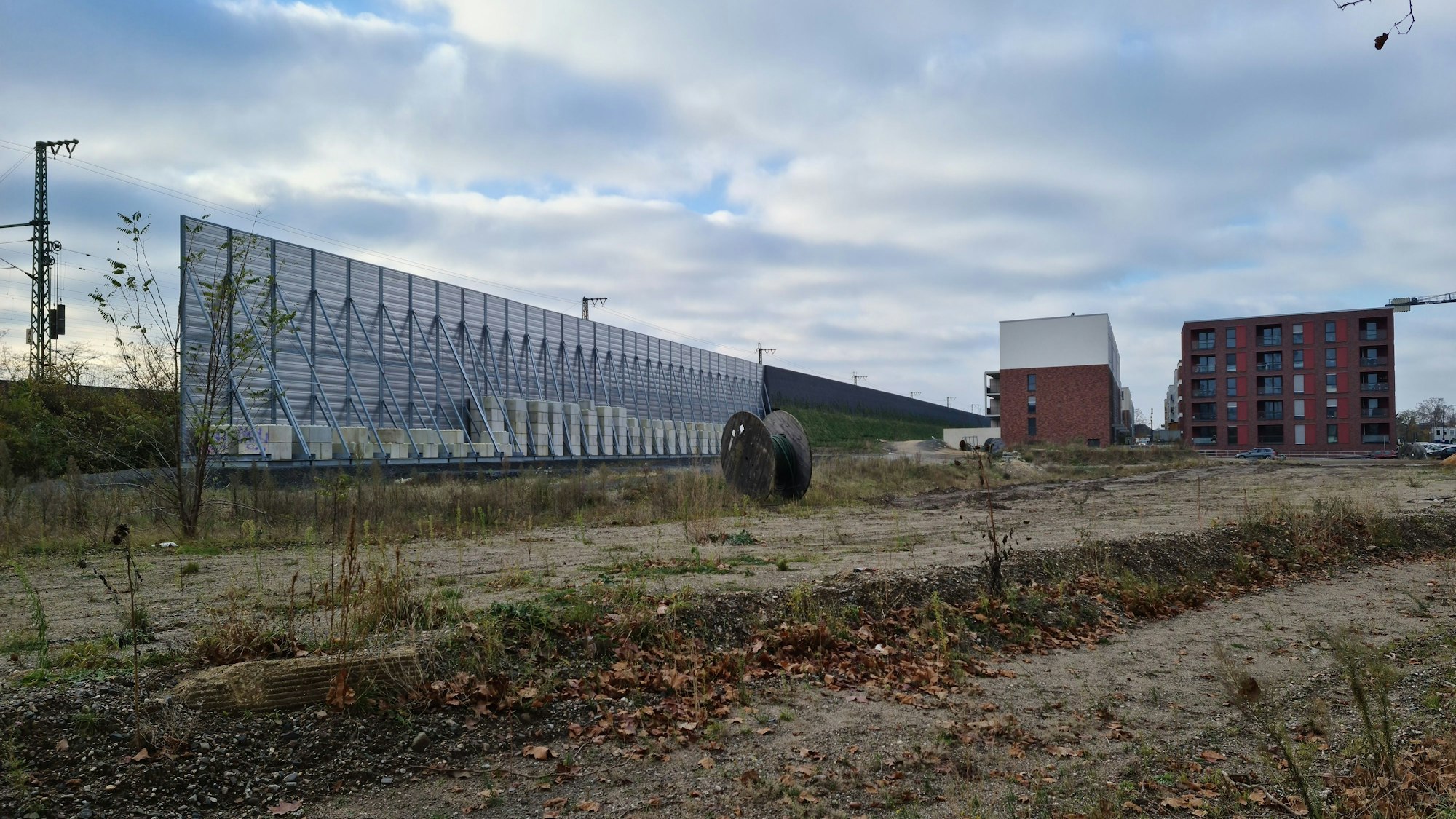 Auf dem Gelände des Alten Güterbahnhofs sieht man viel unbebautes Grundstück, eine sehr hohe Lärmschutzwand an der Bahnlinie und fertig gestellte Wohnblocks im Hintergrund.