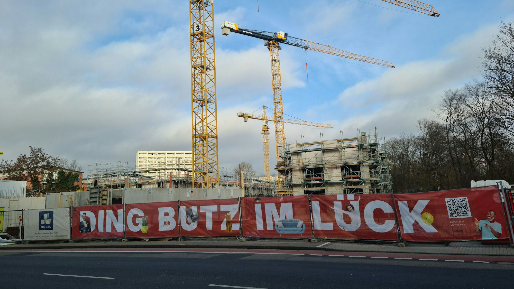 Drei Kräne stehen hinter einem Bauzaun an einer zweispurigen Straße.