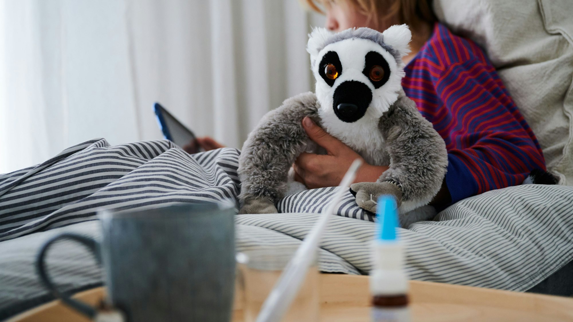 Ein Fieberthermometer, Nasenspray und eine Tasse Tee stehen vor einem Bett, in dem ein Kind mit Kuscheltier sitzt und auf ein Tablet schaut.