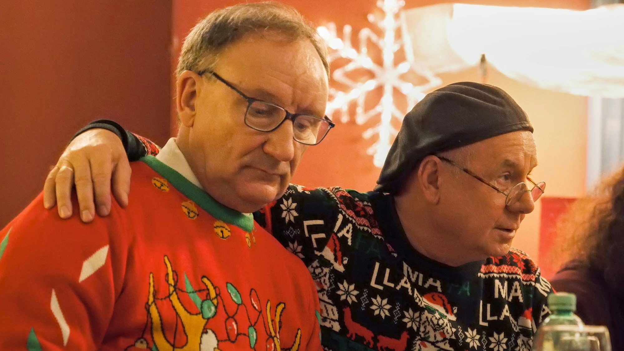 Rainer Bock und Joachim Król sitzen Arm in Arm und tragen Weihnachtspullis.