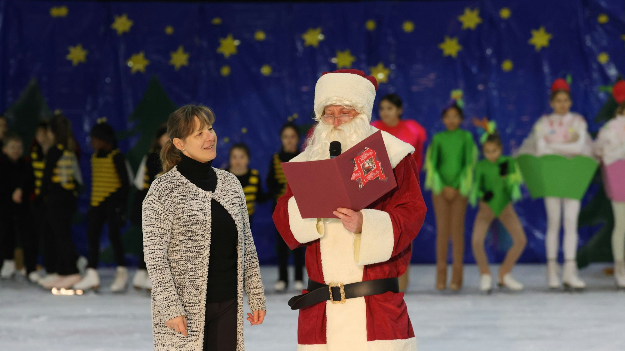 Ein Weihnachtsmann steht neben einer Frau auf einer Eislaufbahn und liest aus seinem Buch vor. Im Hintergrund sind viele verkleidete Eiskunstläuferinnen zu sehen.