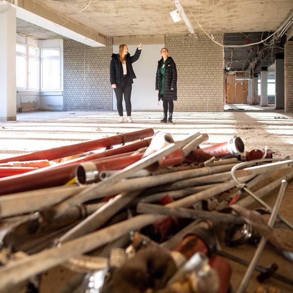 Das Bild zeigt zwei Schülerinnen in dem Rohbau. Kabel hängen von der Decke und Rohre sowie Leitungen liegen auf dem Boden.