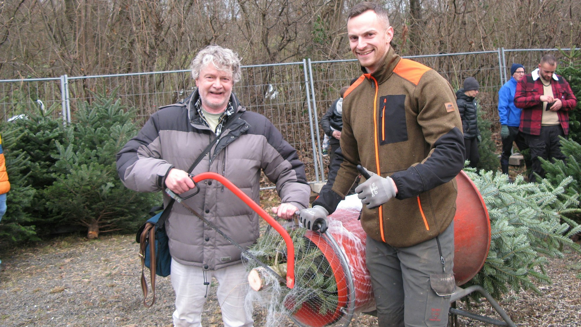 Der Zeitungsreporter mit Säge steht mit dem Weihnachtsbaumverkäufer an der Röhre, in dem der selbst geerntete Christbaum zum Einnetzen steckt.