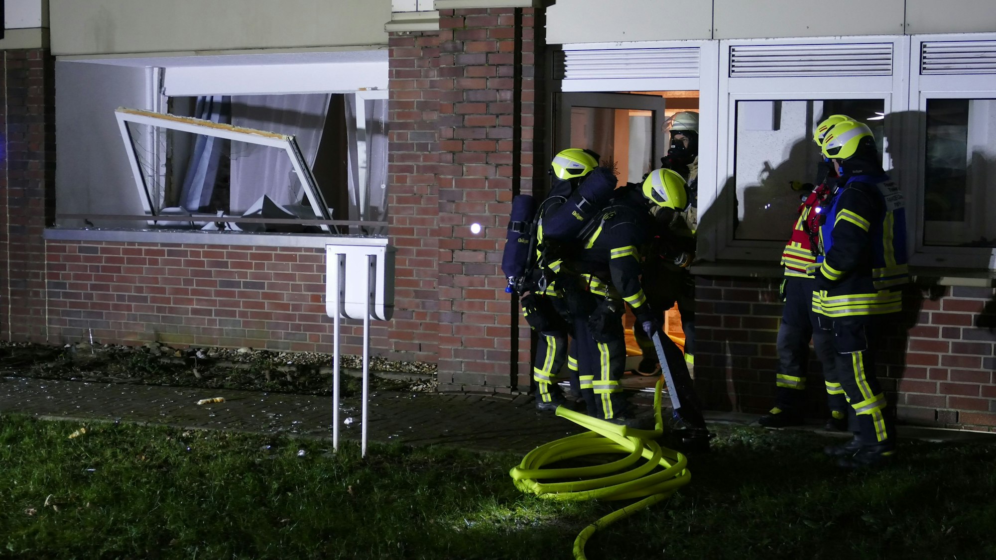 Feuerwehrleute stehen vor einer Wohnung, daneben ist ein beschädigtes Fenster zu sehen.