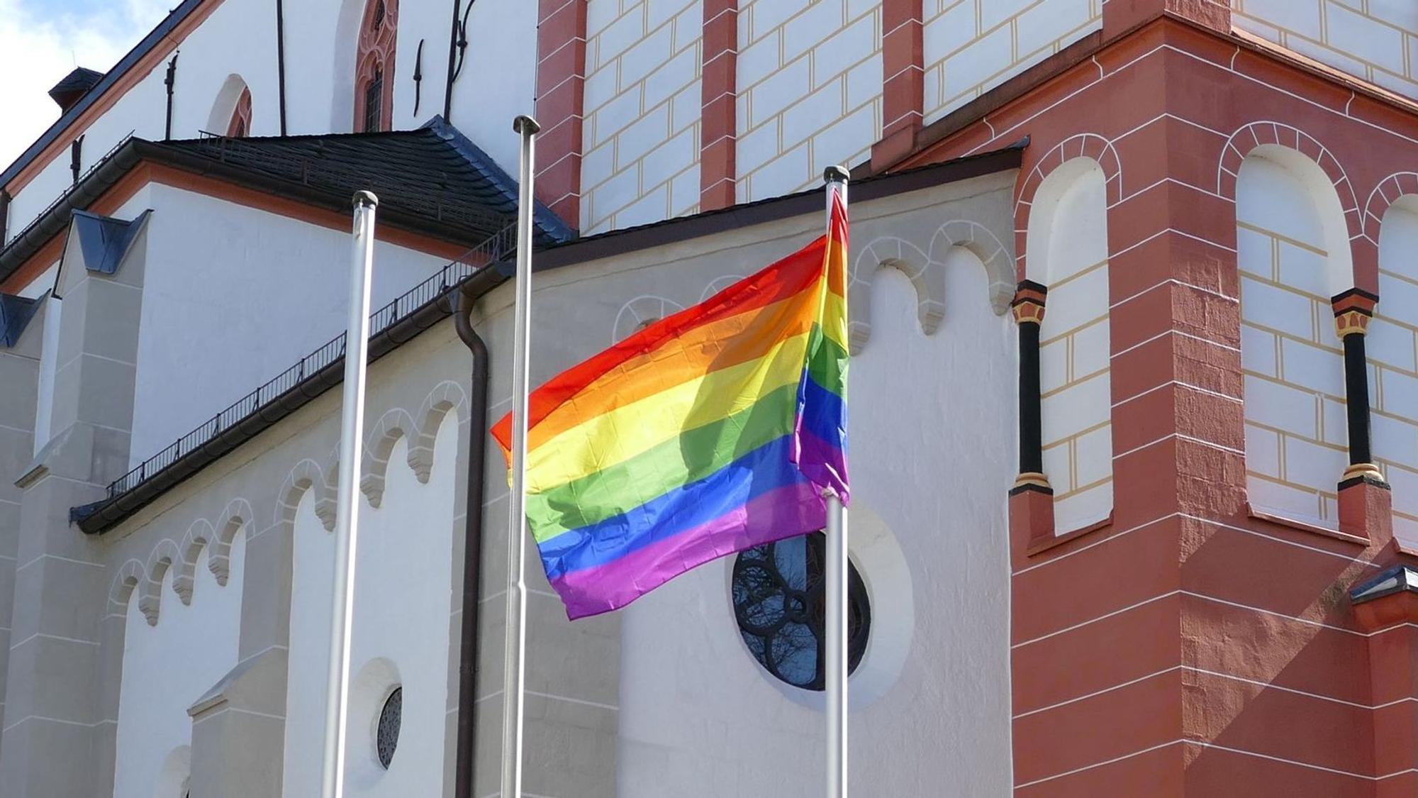 Eine Regenbogenflagge vor einer Kirche mit weiß-ziegelfarbener Fassade
